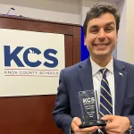 John Clugg holding an award while wearing a jacket and tie in front of a KCS plaque